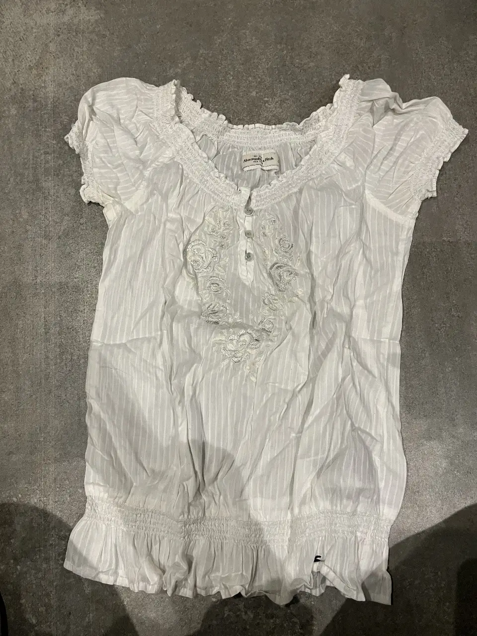 Blusa blanca de Abercrombie & Fitch, con detalles bordados y fruncidos. Ideal para un look casual y fresco.