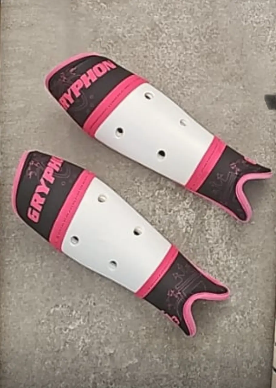 Par de canilleras deportivas de la marca Gryphon, en color blanco con detalles en negro y rosa. Ideales para la práctica de hockey.