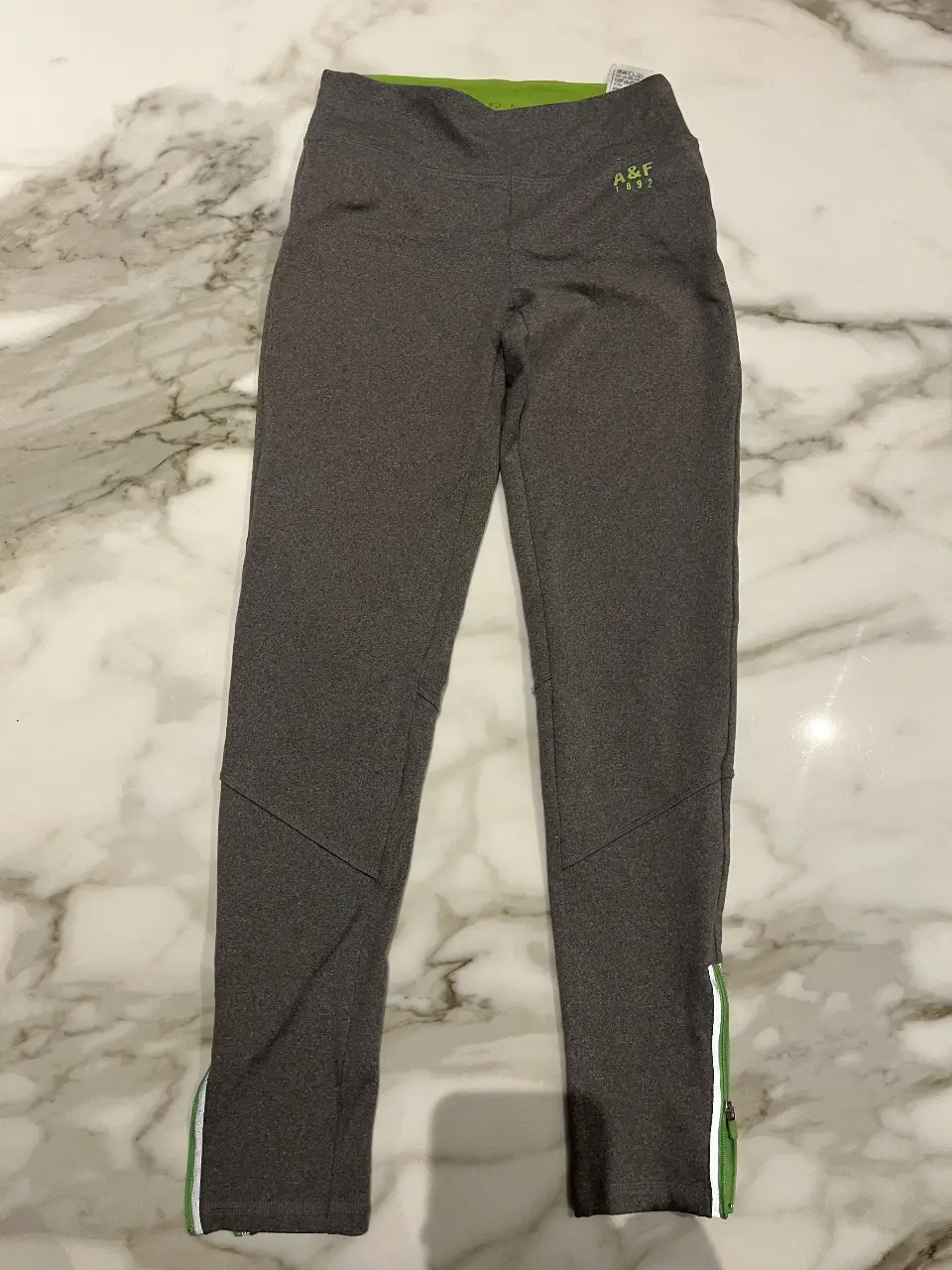 Calzas deportivas grises de mujer con detalles en verde y cierre en los tobillos. Ideal para actividades físicas.