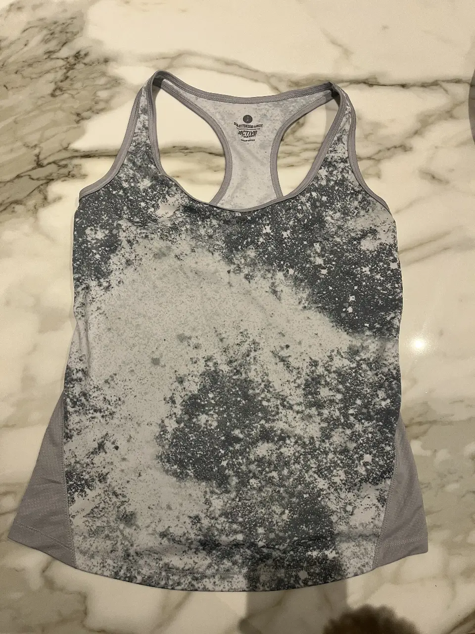 Musculosa deportiva para mujer, con diseño jaspeado en tonos grises y blancos. Ideal para entrenamientos y actividades físicas. Confeccionada con materiales transpirables y cómodos.