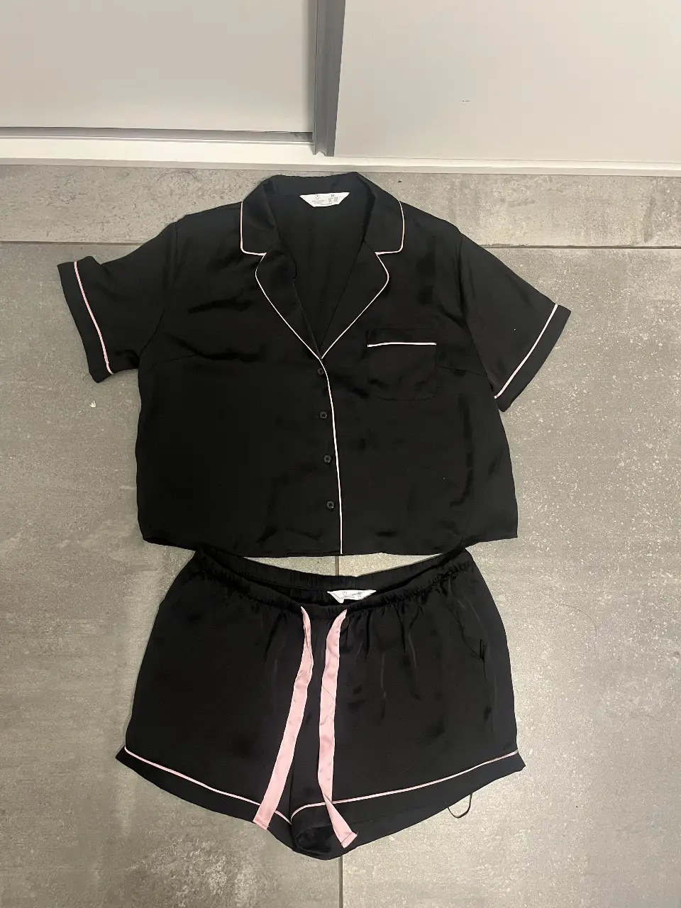 Conjunto de pijama de dos piezas, negro con detalles en rosa. Incluye una camisa de manga corta con botones y un short con cintura elastizada y cordón. Ideal para un descanso cómodo y con estilo.