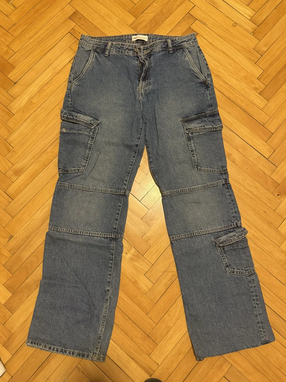 Jeans cargo de denim azul. Poseen múltiples bolsillos con solapa en los laterales de las piernas y en la parte trasera. Cierre y botón en la parte delantera.