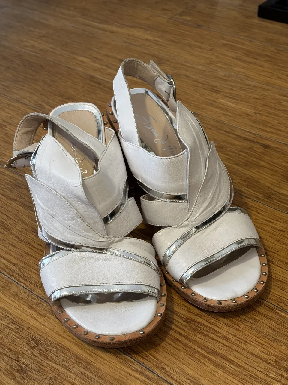 Sandalias de cuero blancas con detalles plateados y taco de madera. Diseño con apliques de hojas y tachas en la suela. Ideales para un look elegante y moderno.