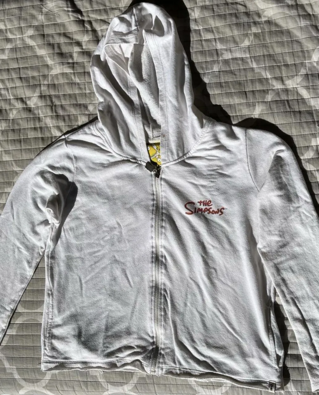 Buzo con cierre blanco de The Simpsons, con capucha y estampado de Bart Simpson en la espalda. Ideal para un look casual y cómodo.