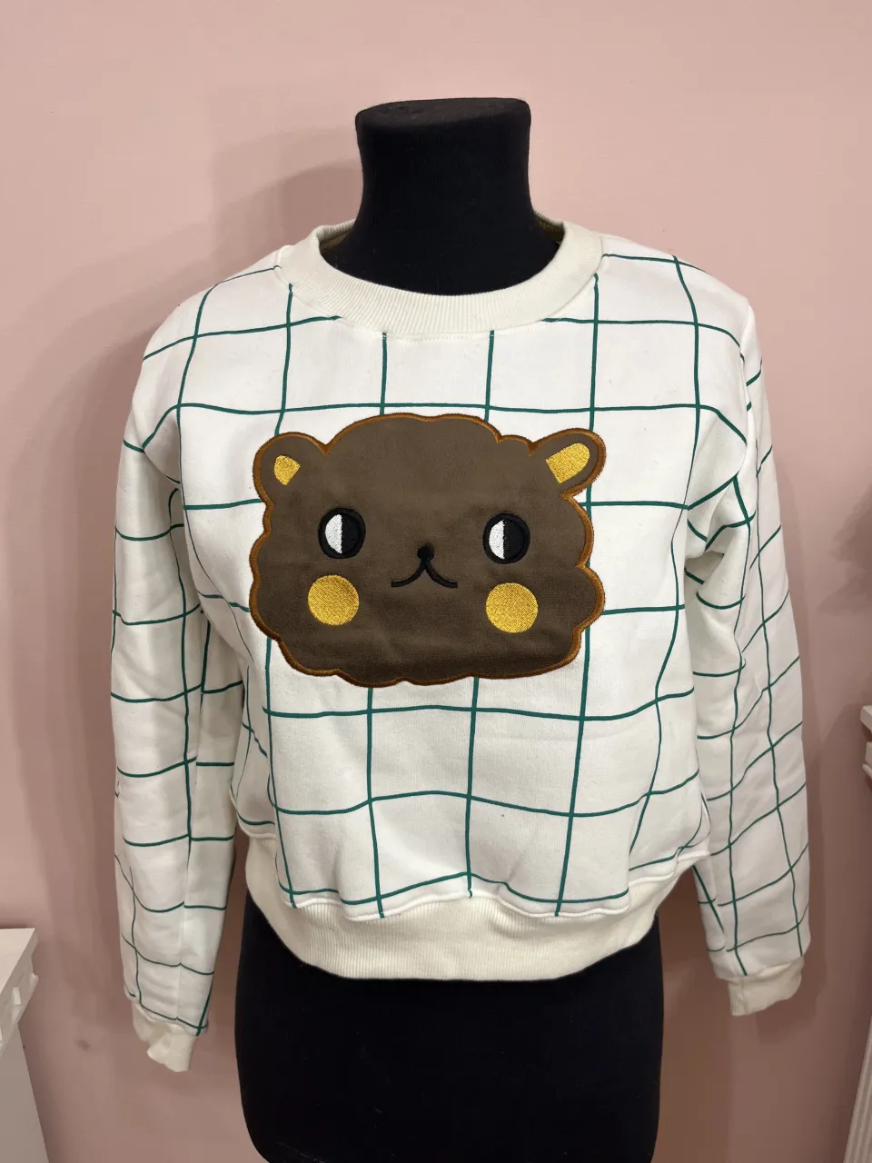 Buzo blanco con estampa de cuadrícula verde y un aplique de oso marrón en el frente. Tiene parches de gamuza en los codos.