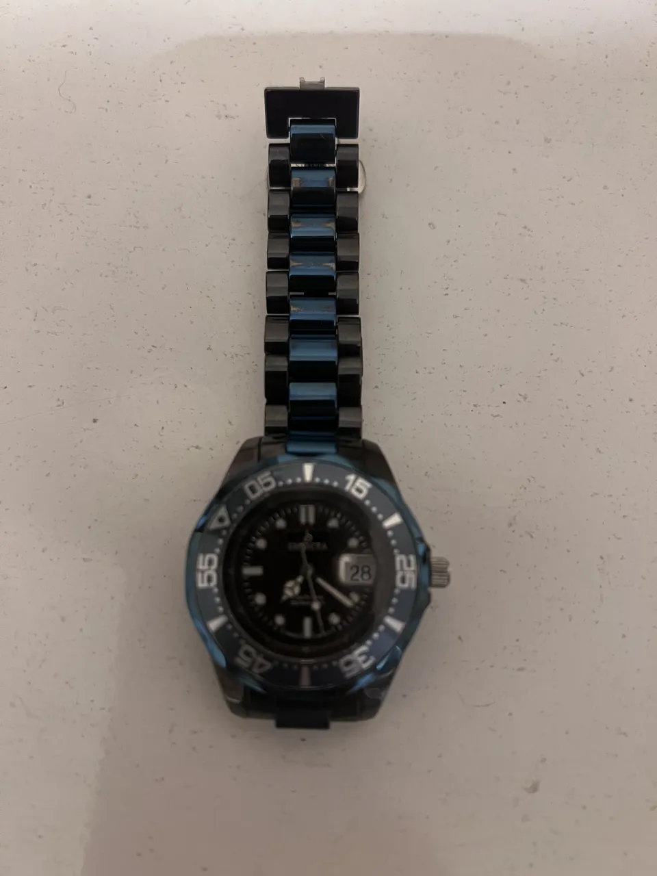 Reloj de pulsera con malla metálica y bisel azul. La esfera es negra con detalles plateados y números blancos. Tiene fechador.