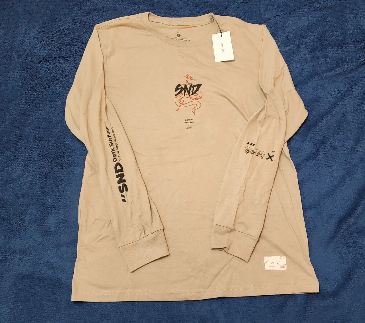 Remera manga larga de color crudo con estampado "SND" y diseño de serpiente en el frente. Detalles de texto en las mangas y en la parte trasera. Ideal para un look casual y urbano.