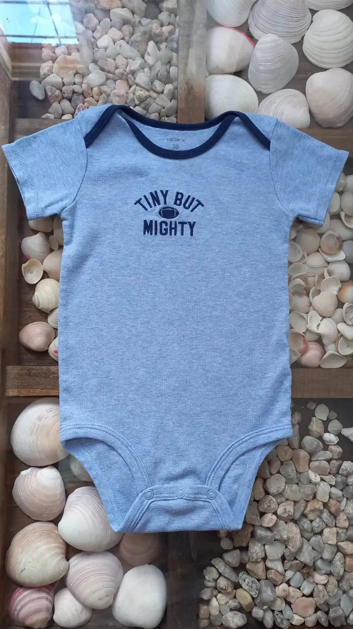 Body de bebé manga corta, color azul claro, con estampa "TINY BUT MIGHTY" y un dibujo de pelota de fútbol americano. Cuello y puños con borde azul marino.