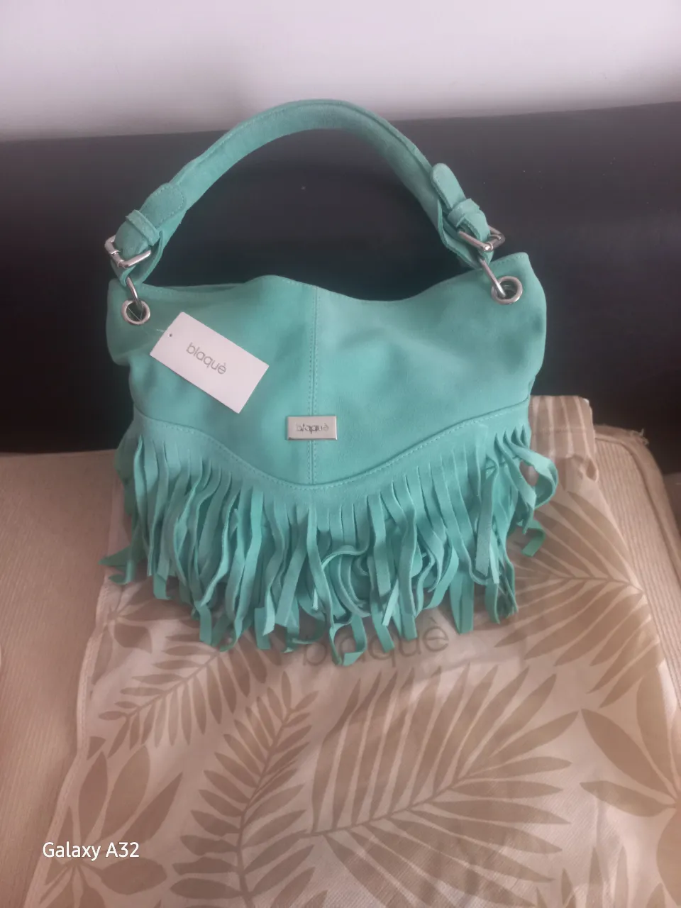Cartera bandolera de gamuza color verde agua  con flecos en la parte inferior. Tiene un asa corta para llevar en la mano y una etiqueta con el logo de la marca.
