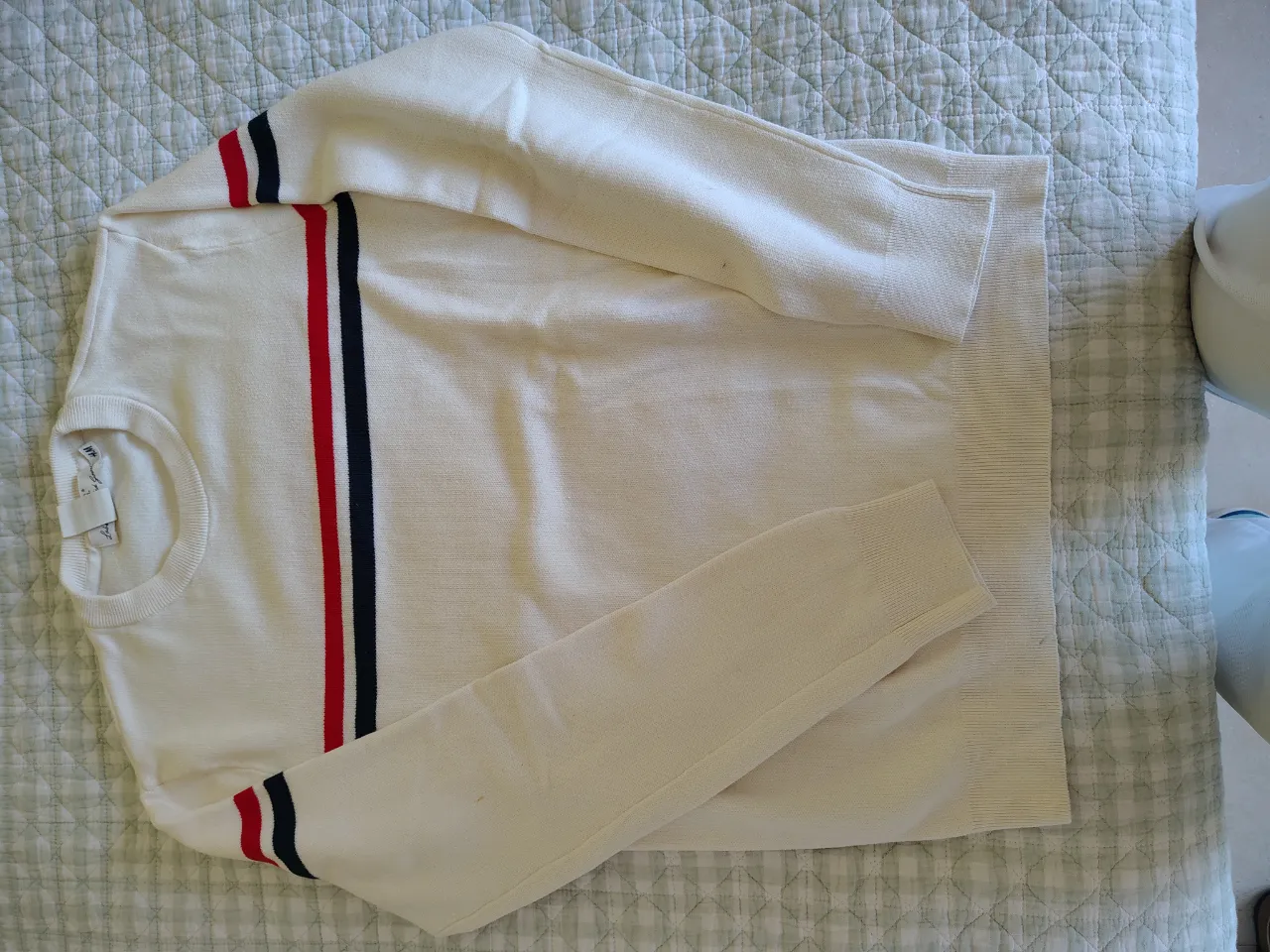 Sweater blanco con cuello redondo y mangas largas. Presenta dos franjas verticales en el frente, una roja y una azul marino, y detalles de las mismas franjas en los puños de las mangas. También tiene franjas rojas y azul marino en los hombros.