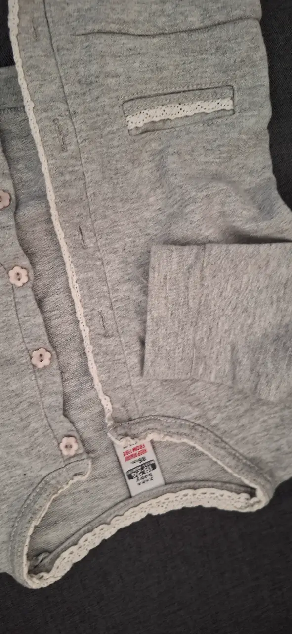 Cardigan gris de bebé con detalles de encaje blanco. Cierre con botones florales. Ideal para mantener a tu bebé abrigado con estilo.