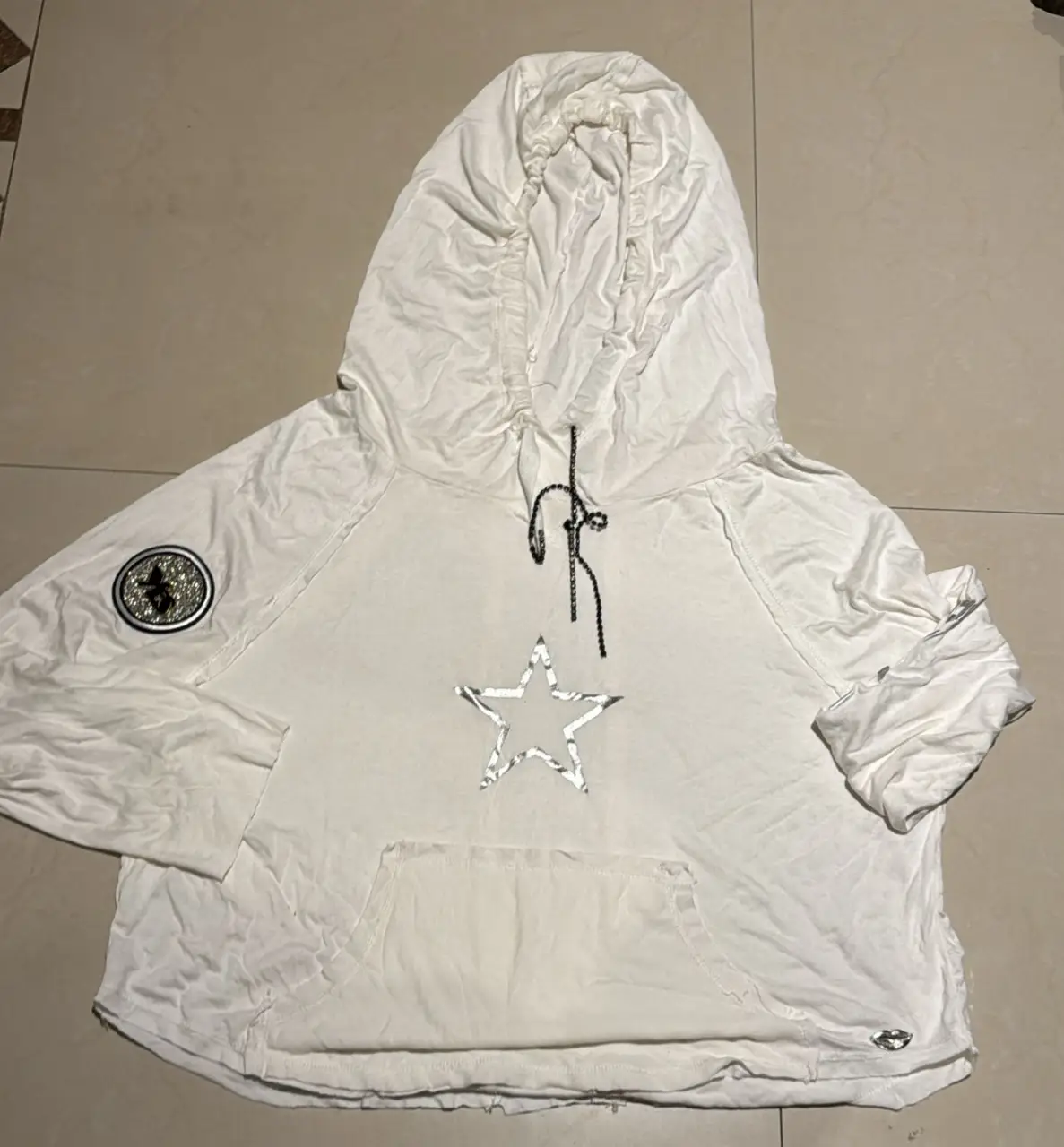 Buzo canguro blanco con capucha y detalle de estrella plateada. Cuenta con un parche con brillos y detalles en las mangas. Ideal para un look casual y cómodo.