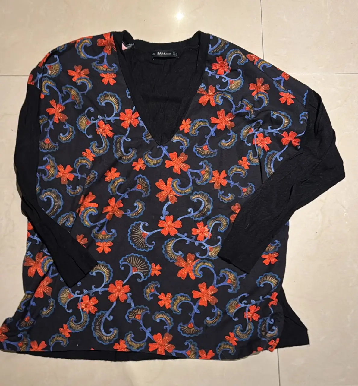 Blusa de manga larga con estampado floral en tonos naranja y azul sobre fondo negro. Cuello en V y mangas negras lisas. Ideal para un look casual con un toque bohemio.