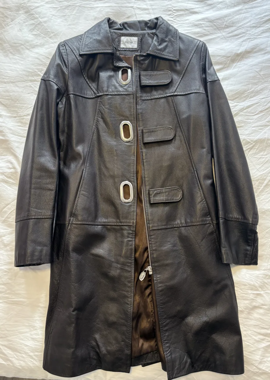 Abrigo de cuero marrón oscuro, largo hasta arriba de la rodilla, con cuello solapa y detalles de costura. Posee botones grandes y un cierre metálico. Ideal para un look elegante y sofisticado.