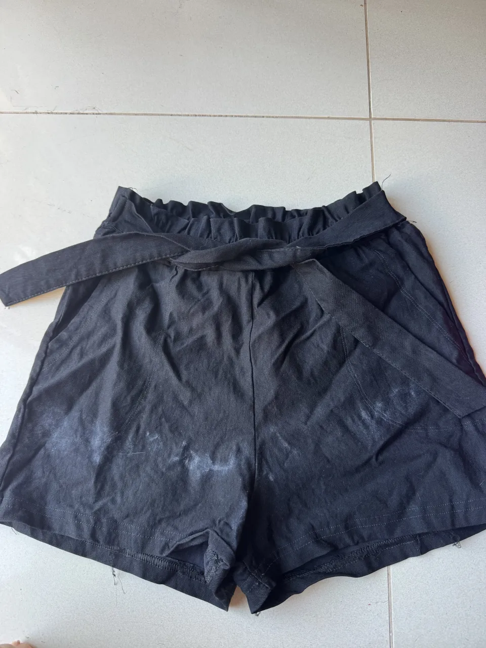 Shorts negros con cinturón de tela y elástico en la cintura. Tienen detalles de desgaste o manchas blancas en la parte delantera.