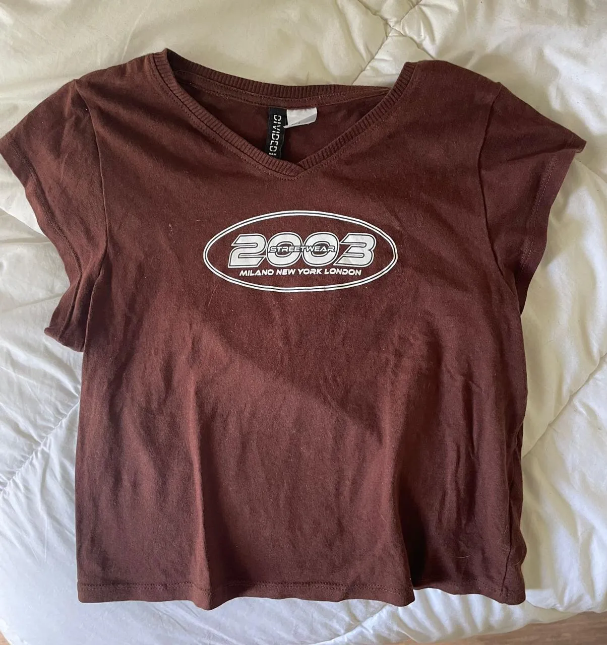 Remera marrón de manga corta con cuello en V y estampa "2003 STREETWEAR MILANO NEW YORK LONDON" en blanco.