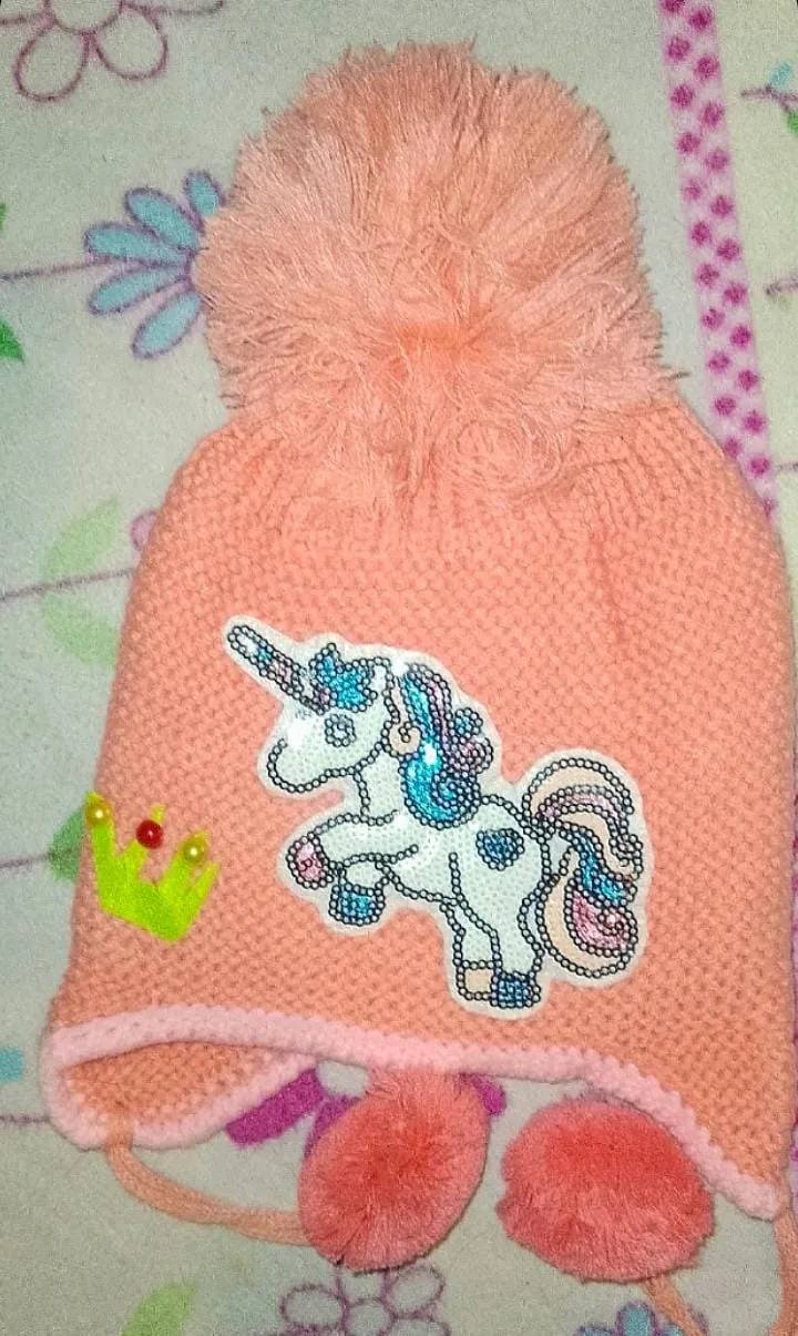 Gorro tejido de lana color rosa con pompón grande en la parte superior. Tiene un aplique de unicornio con lentejuelas y una corona con detalles brillantes. Incluye orejeras con pompones colgantes.