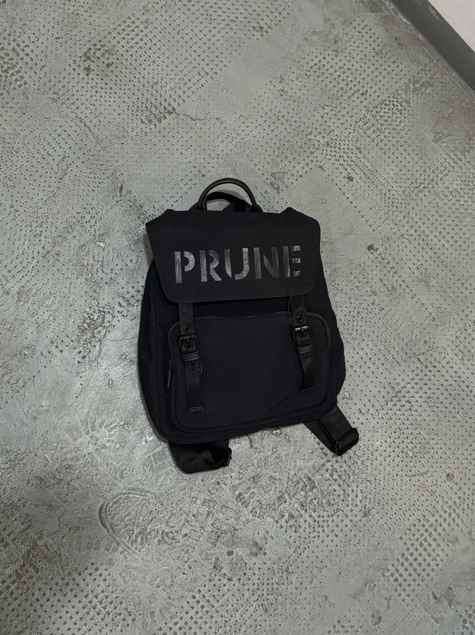 Mochila negra PRUNE, con solapa y hebillas. Ideal para llevar tus cosas con estilo y comodidad.