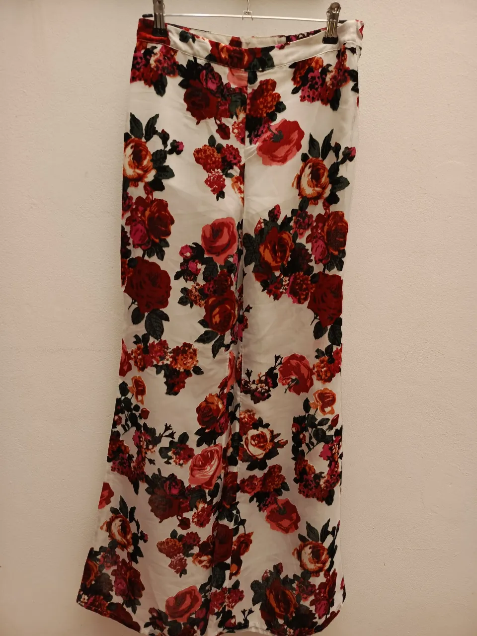 Pantalón blanco con estampado floral en tonos rojos y rosas. Diseño de flores grandes y pequeñas distribuidas por toda la prenda.