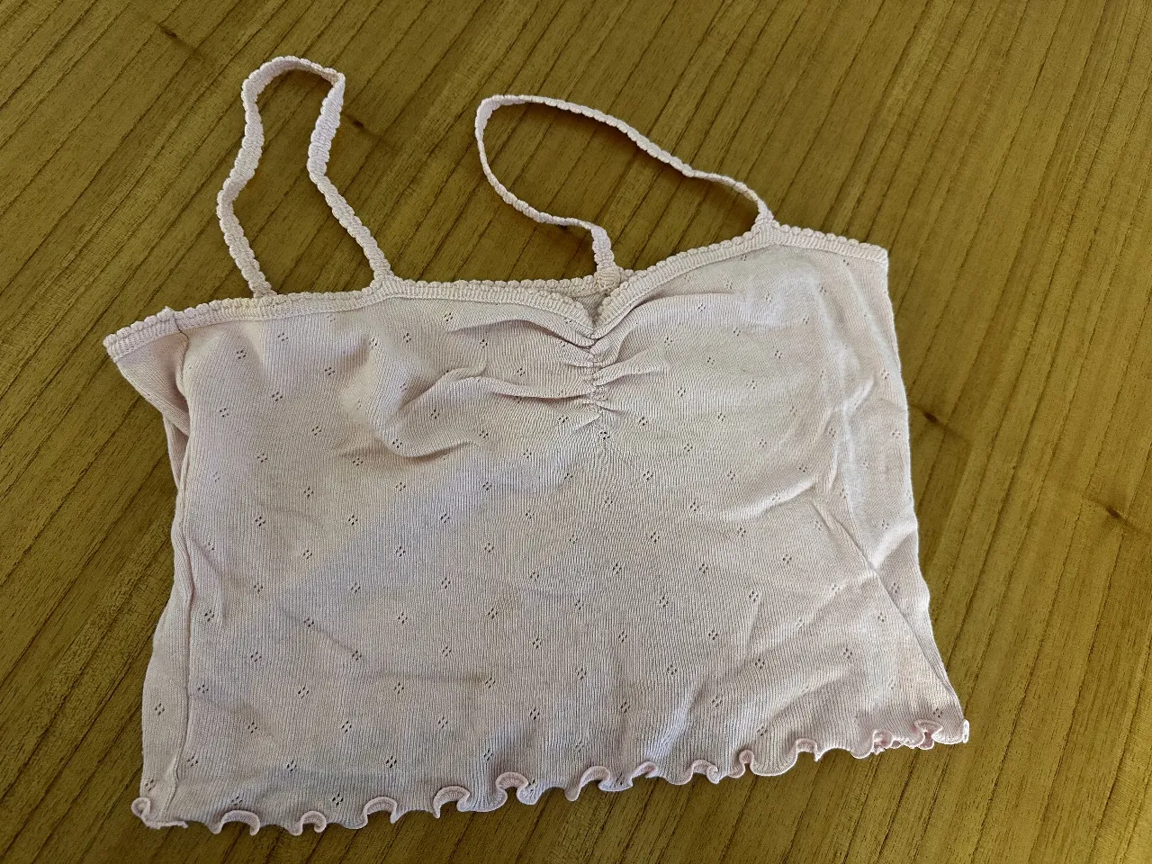 Top de mujer de color rosa claro, con breteles finos y detalle fruncido en el centro del busto. Diseño delicado con bordes ondulados y pequeños detalles perforados en la tela.