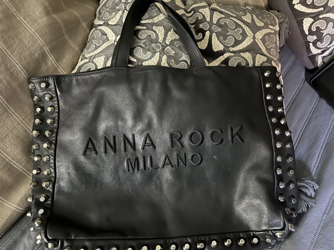 Bolso tote de cuero negro, súper suave, marca italiana. Tachas plateadas metálicas. Cierre con broche imantado y bolsillos internos, uno con cierre. En el frente  logo de marca "ANNA ROCK MILANO", en relieve. 