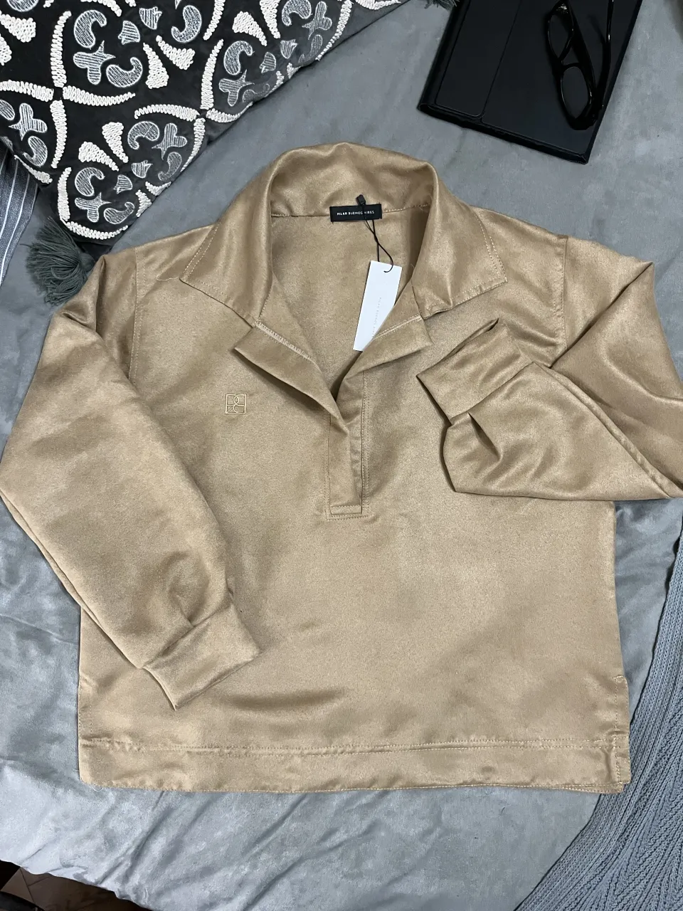 Remera polo (estilo buzo liviano) de escote abierto y profundo, color camel. Manga larga con puños marcados en el mismo género. Posee detalle bordado de logo de la marca en el frente. Corte recto. Ruedo ancho y tajos laterales. 