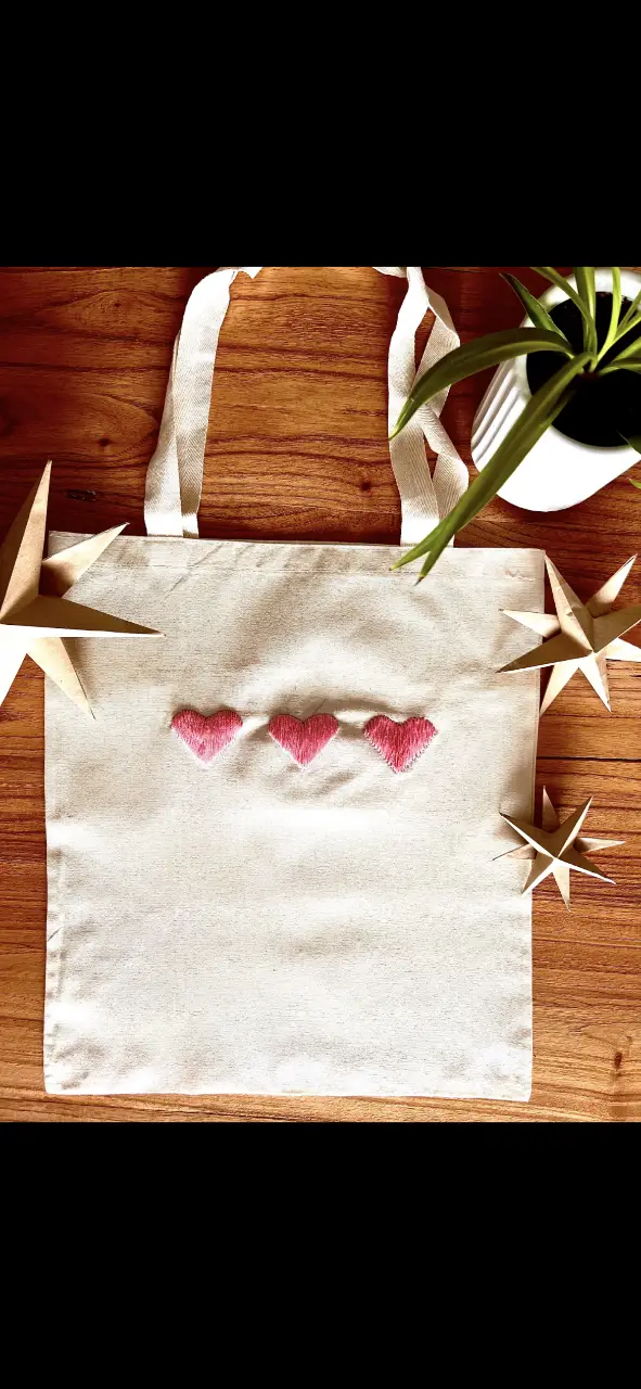 Bolso tote de tela cruda con tres corazones bordados en rosa. Ideal para llevar tus cosas con estilo y un toque artesanal.