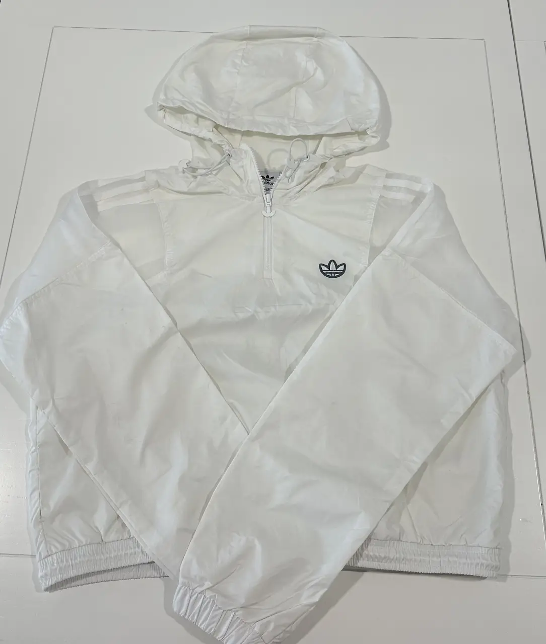 Campera rompevientos Adidas blanca con capucha y cierre. Ideal para actividades deportivas o uso casual. Posee el logo de la marca en el pecho y en la espalda.