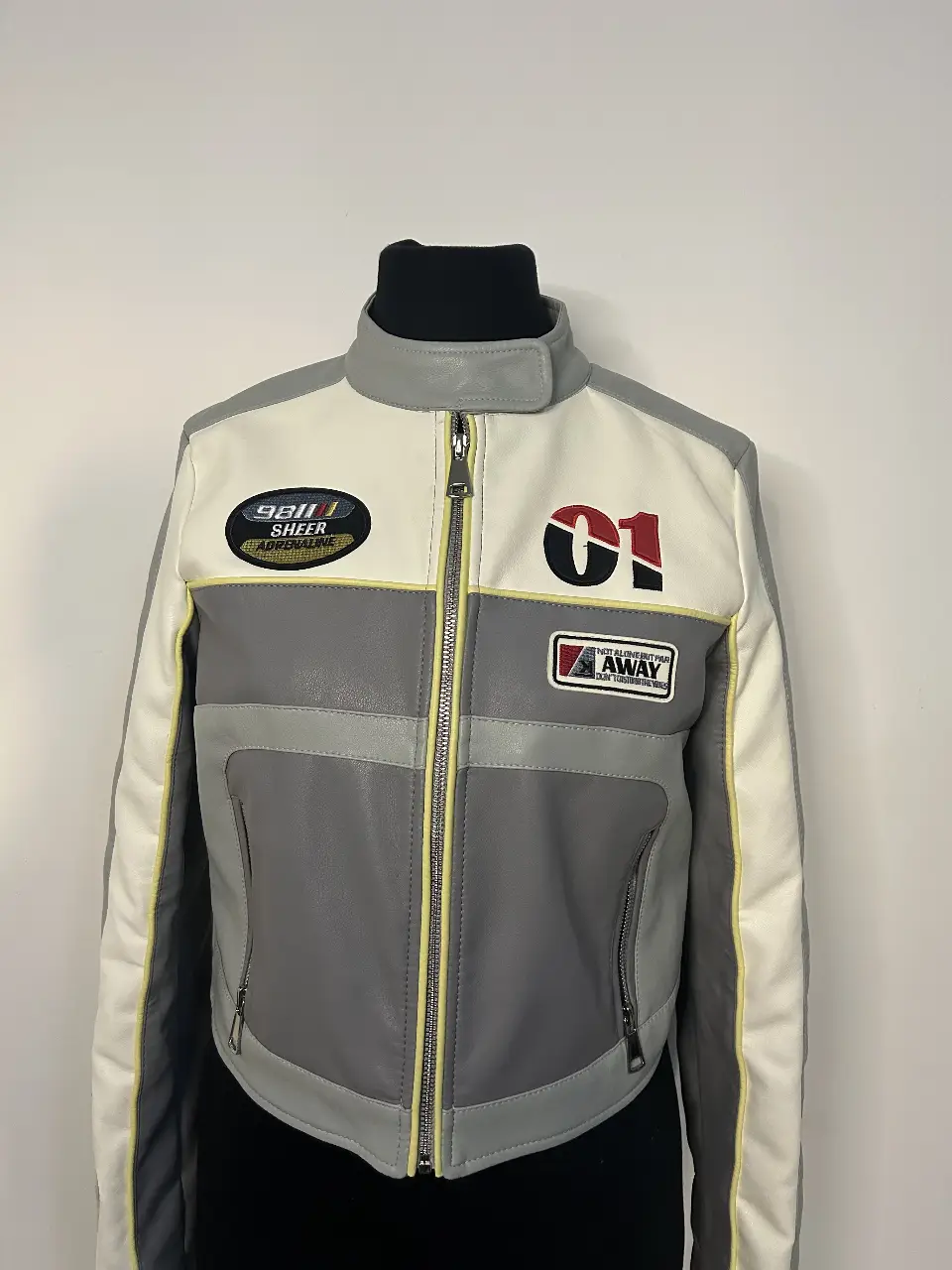 Chaqueta estilo biker de cuero sintético en tonos gris y blanco con detalles en amarillo. Diseño con parches y detalles deportivos.