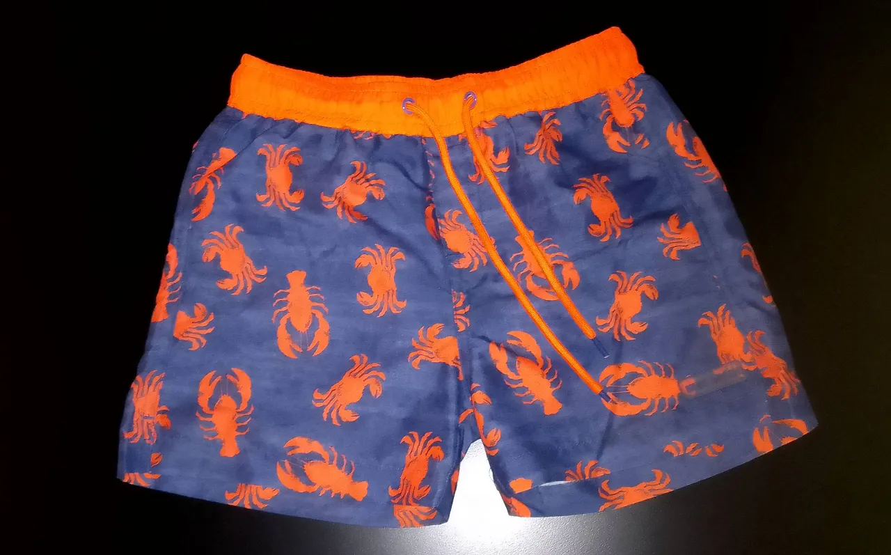 Short de baño para niño, con estampado de langostas en color naranja sobre fondo azul. Cintura elastizada con cordón ajustable. Ideal para la playa o pileta.