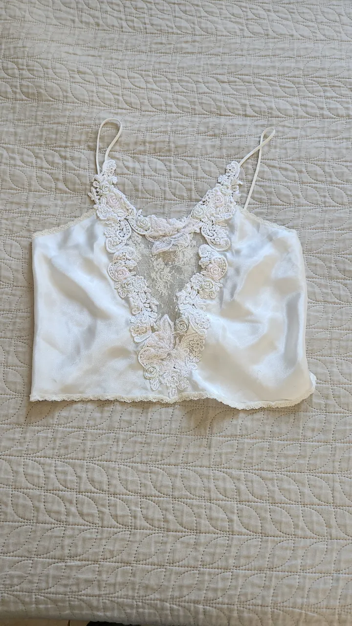 Top de satén blanco con detalles de encaje floral y lazo en el escote. Breteles finos y terminación de encaje en el borde inferior.