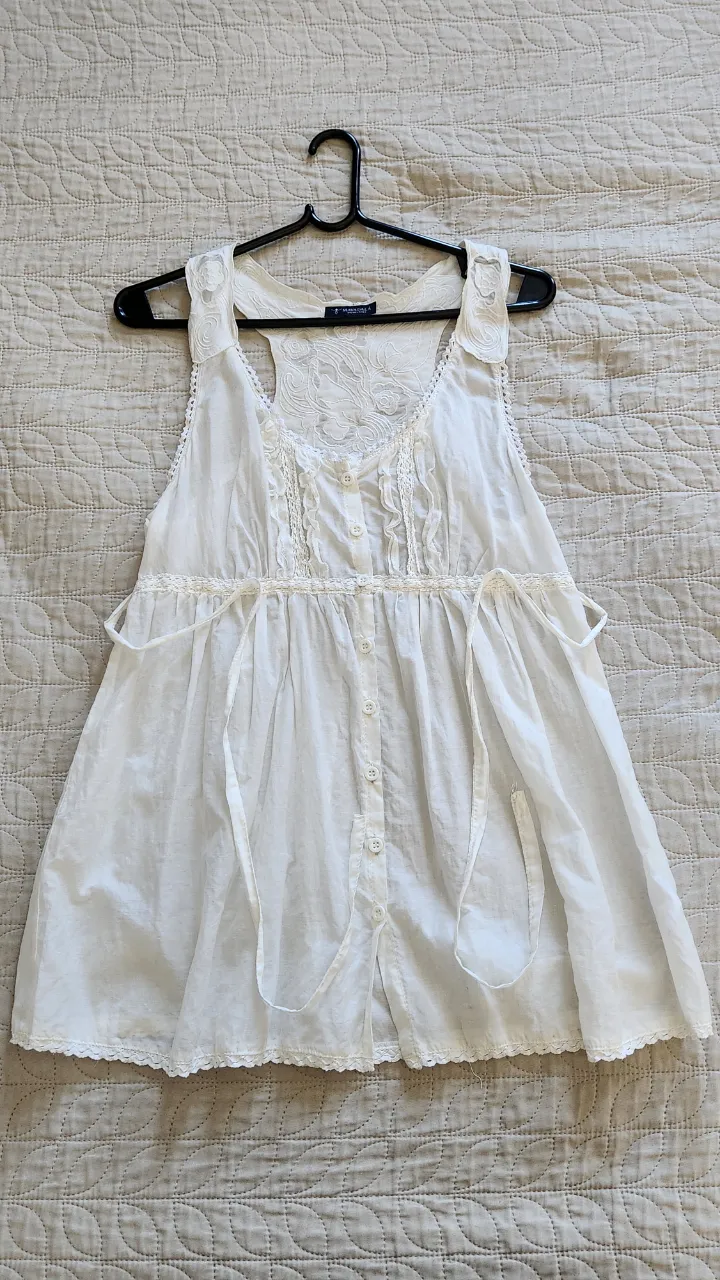 Musculosa blanca con detalles de encaje y bordados en la parte superior. Posee botones al frente y lazos a los costados para ajustar.