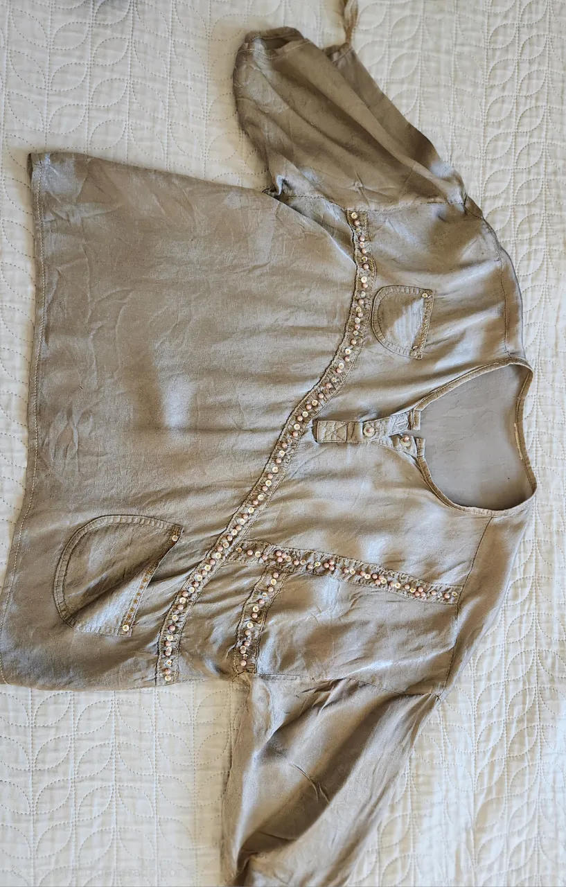 Remera de manga larga color beige con detalles de cuentas y lentejuelas en el frente. Posee dos bolsillos pequeños en la parte superior y un par de bolsillos más grandes en la parte inferior. Tiene botones en la parte superior del escote.