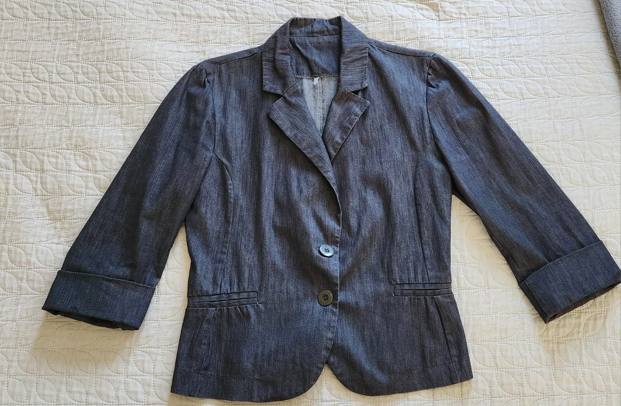 Blazer de tela denim gris oscuro, con cuello solapa, un botón de cierre y bolsillos frontales. Las mangas son 3/4 con puños doblados.