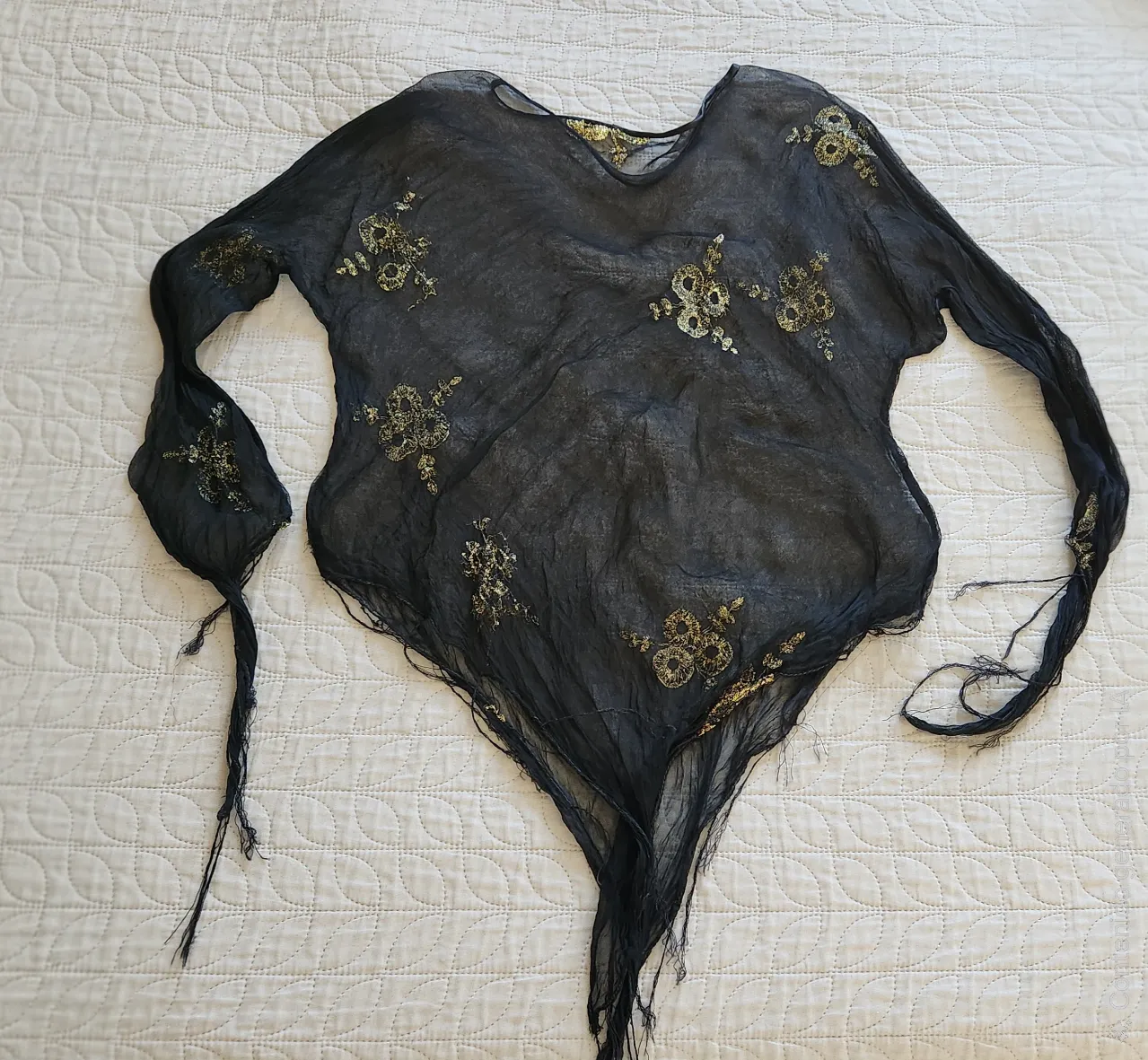 Prenda de tela fina negra con apliques dorados bordados en forma de flores y hojas. Posee mangas largas y terminación deshilachada en puños y ruedo.