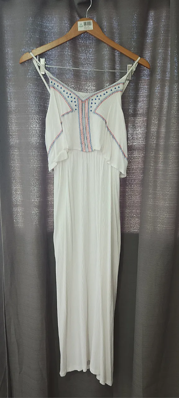 Vestido blanco largo con tiras finas. Posee un detalle bordado en la parte superior del busto con hilos de colores rojo, azul y blanco. La parte superior del busto es suelta y fruncida, y se une a la falda a la altura de la cintura con un elástico.
