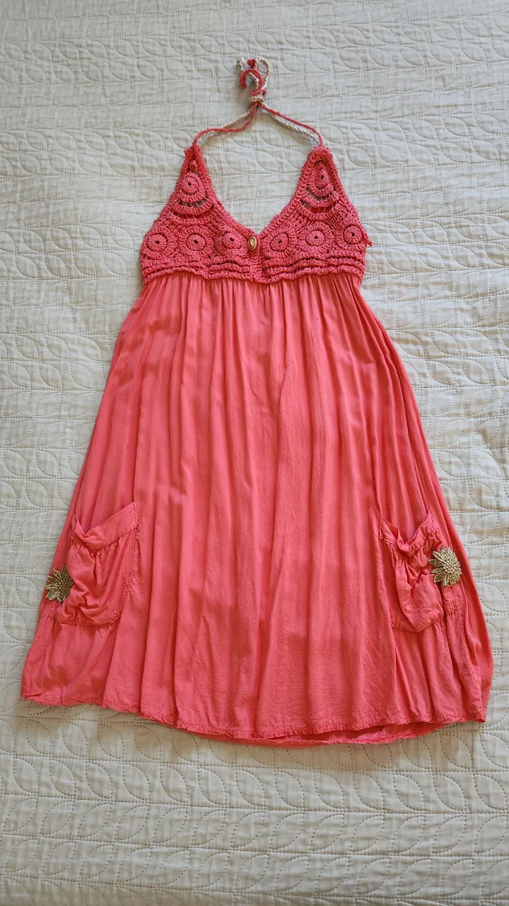 Vestido corto de color coral con detalles de crochet en la parte superior y tiras para atar en el cuello. Posee dos bolsillos delanteros con apliques de flores y detalles dorados.