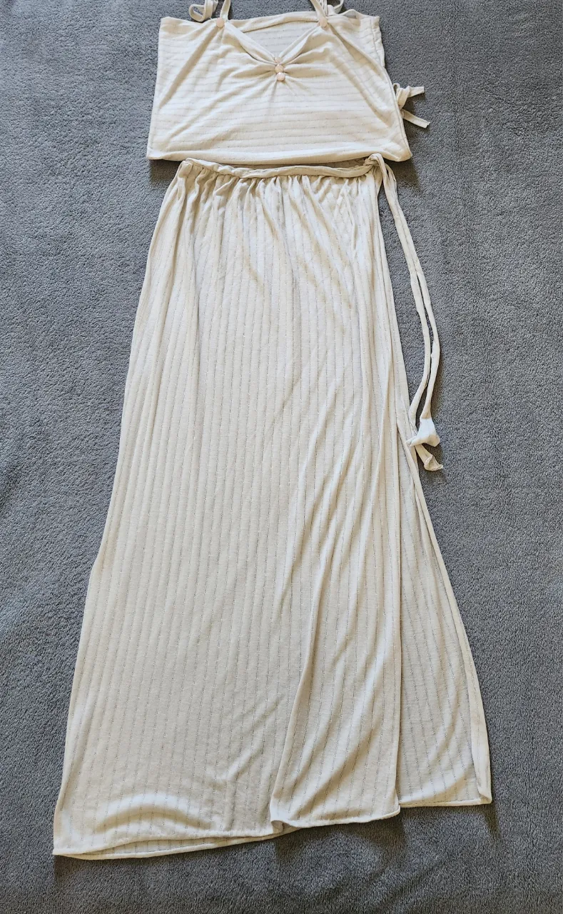 Conjunto de dos piezas compuesto por un top y una falda larga. El top es de breteles finos y tiene un detalle de fruncido con dos botones en el frente. La falda es larga, con cintura elastizada y una abertura lateral. Ambas piezas son de tela rayada fina.