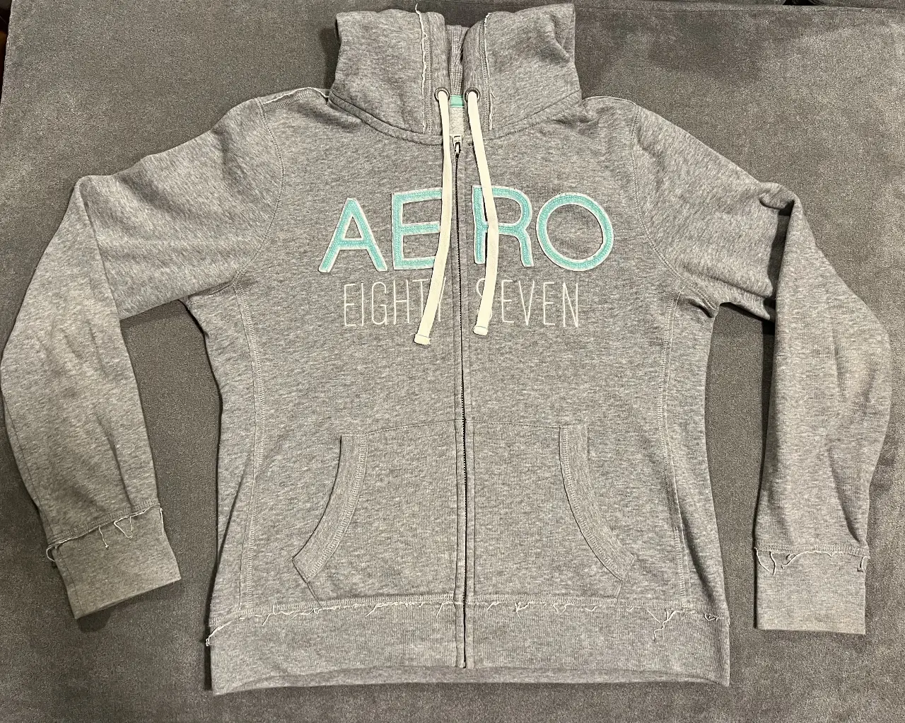 Buzo con cierre gris de la marca Aeropostale, con capucha y el logo "AERO EIGHTY SEVEN" en color verde agua. Ideal para un look casual y cómodo.