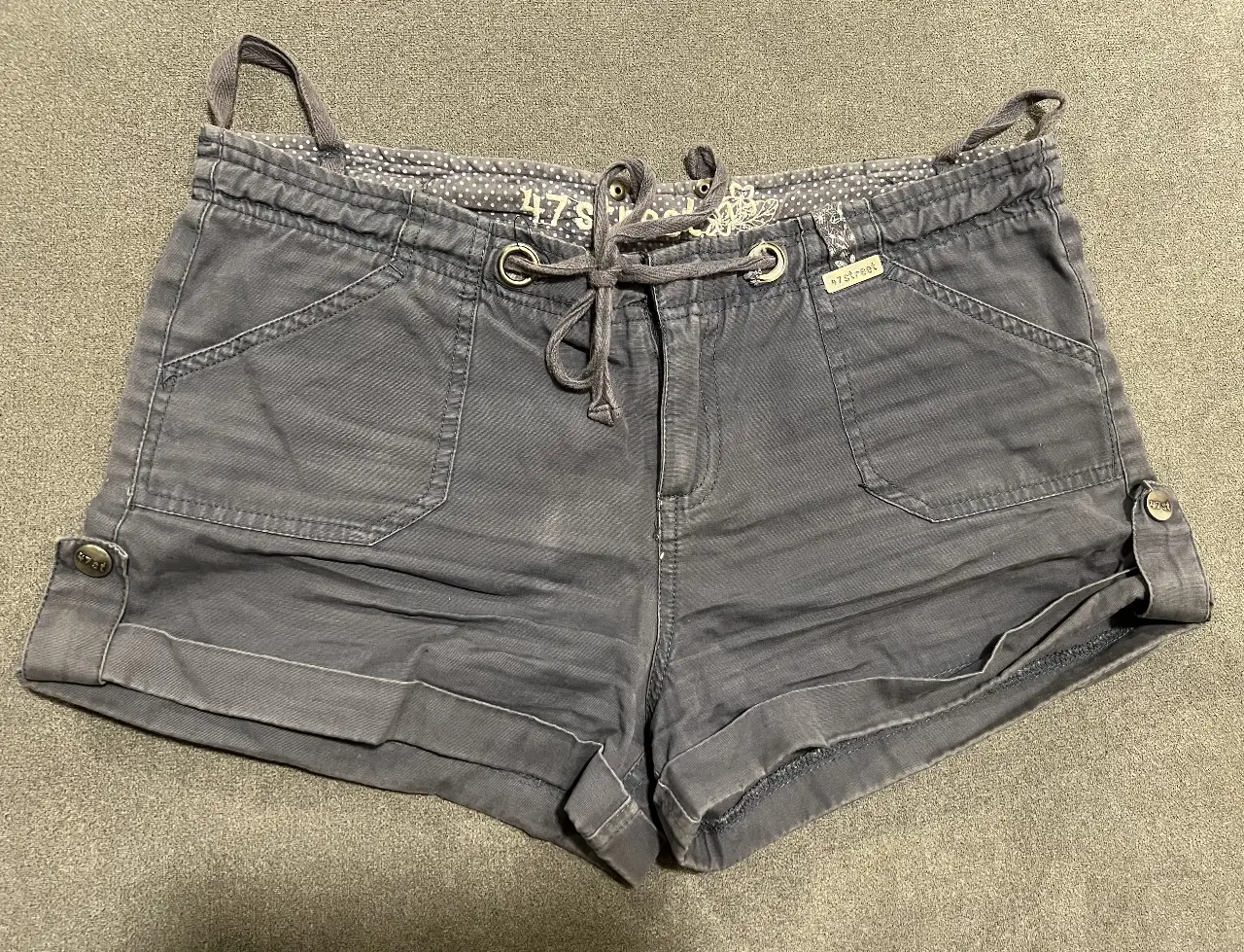 Short de jean gris oscuro de mujer, con detalles de bolsillos delanteros y traseros, ajuste con cordón y botones laterales. Ideal para un look casual y cómodo.