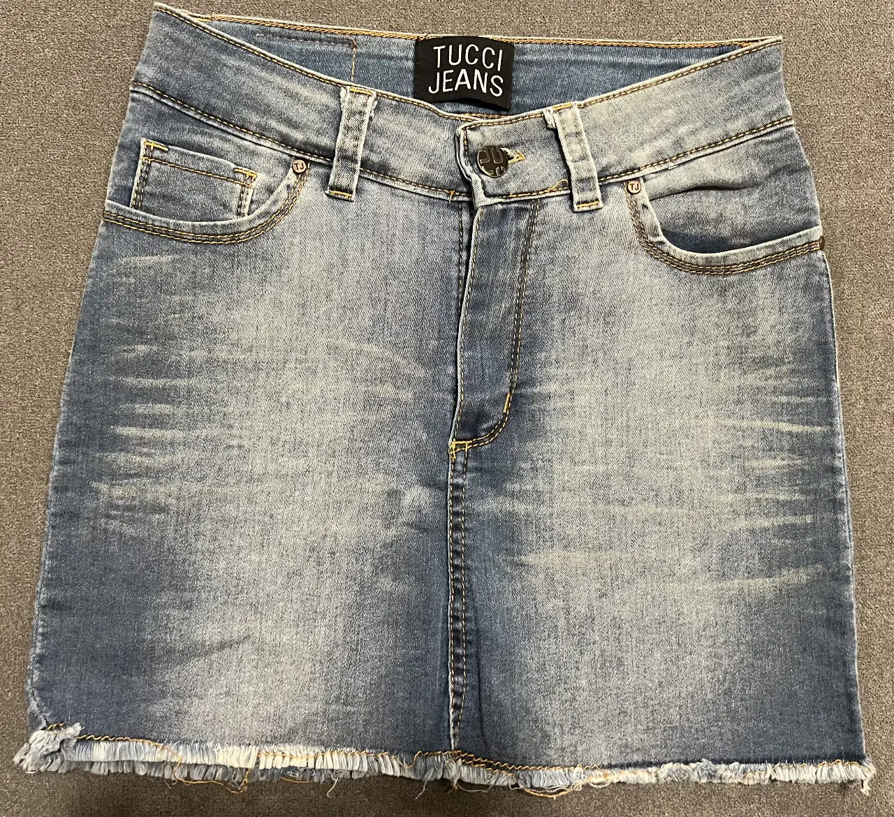 Minifalda de jean azul, con detalles de desgaste y flecos en el borde inferior. Diseño clásico de cinco bolsillos y cierre con botón y cremallera. Ideal para un look casual y moderno.
