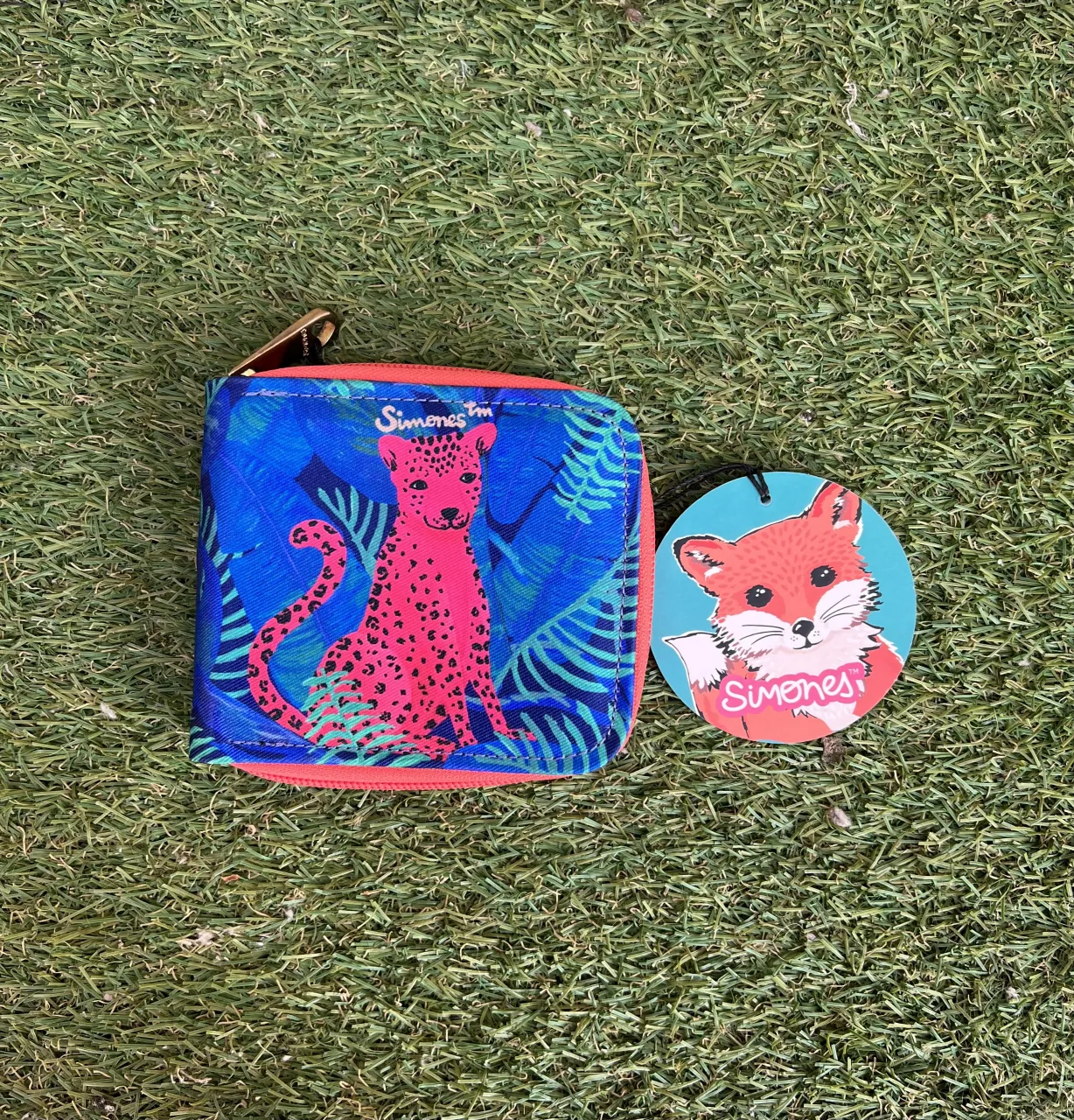 Billetera con estampa de leopardo rosa y hojas tropicales en fondo azul. Cierre de cremallera naranja. Incluye un llavero con forma de zorro.