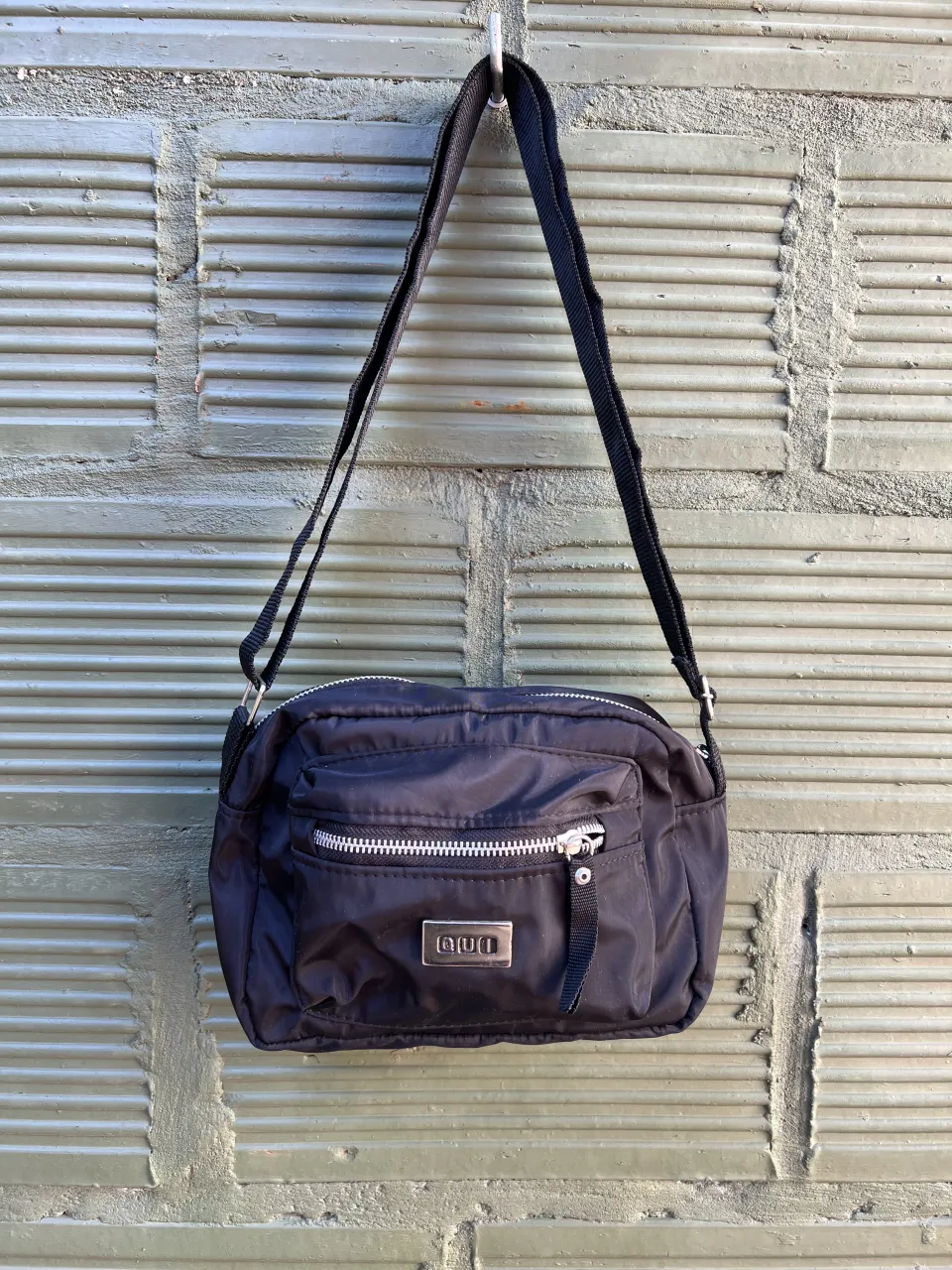 Cartera negra de tela con correa ajustable. Posee un bolsillo frontal con cierre metálico y un bolsillo interno.