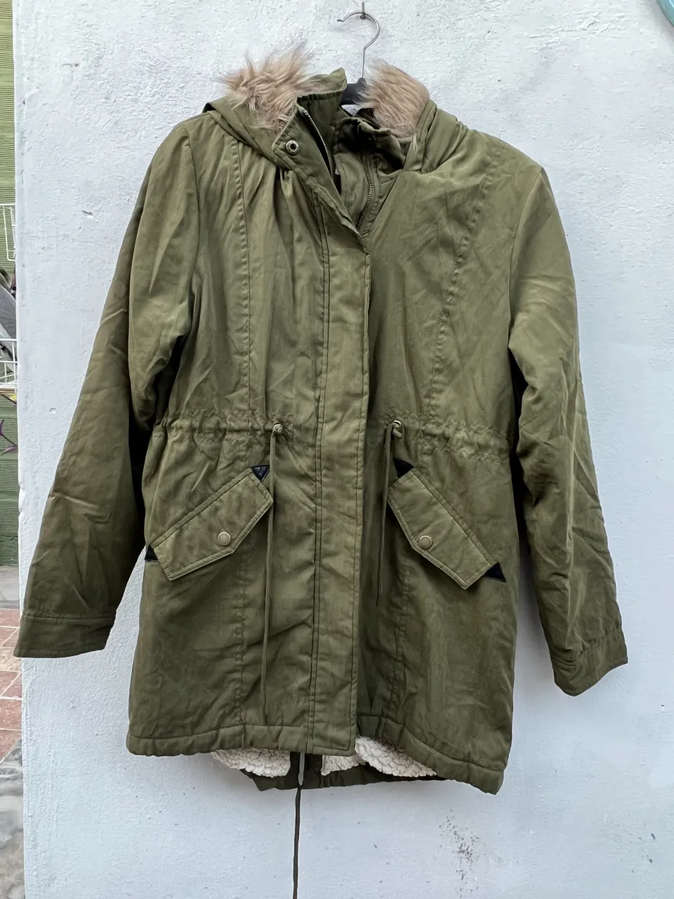 Parka verde con capucha forrada en piel sintética. Posee cordón de ajuste en la cintura y bolsillos con solapa. El interior está forrado con corderito color crudo.