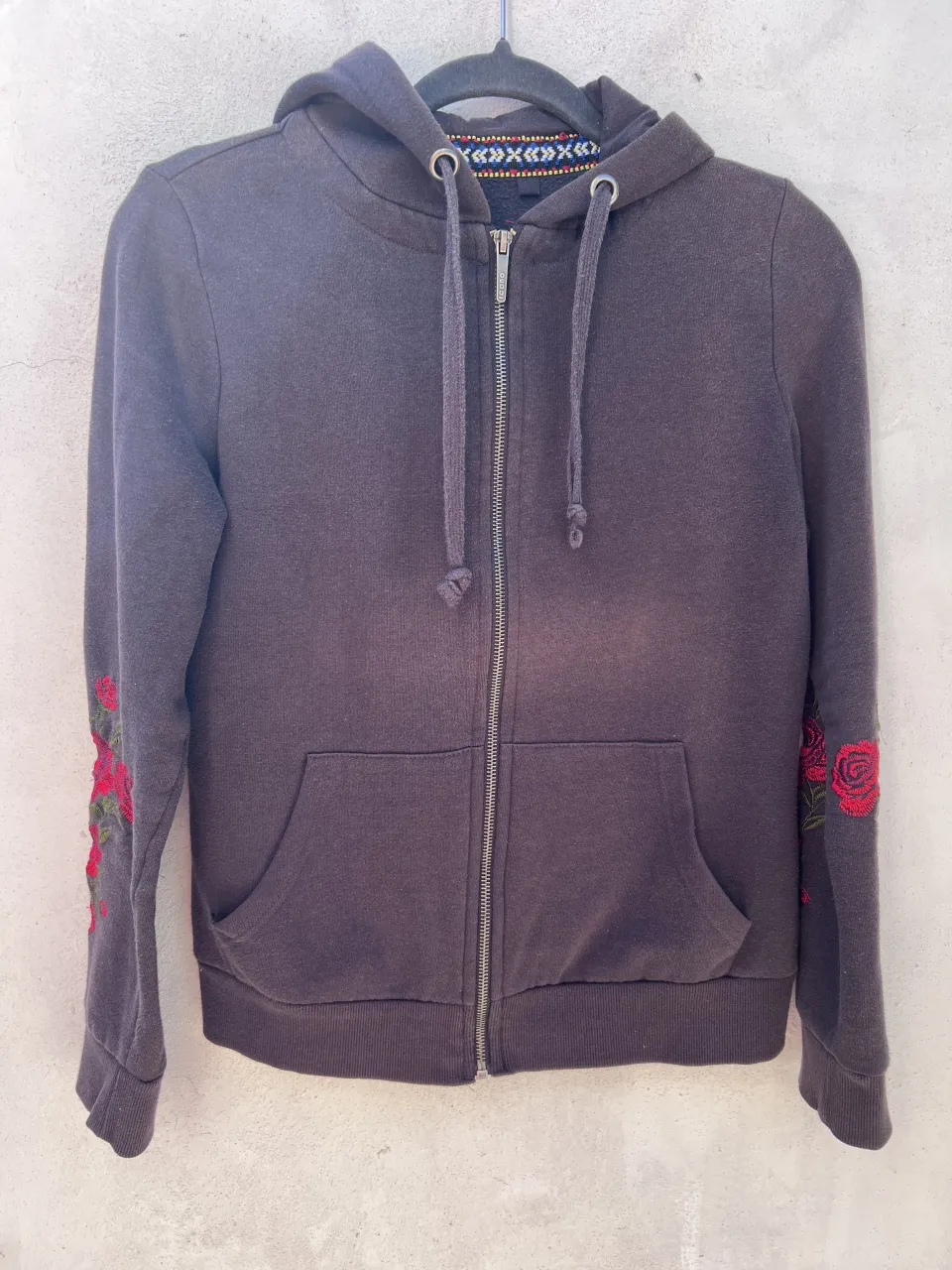 Buzo gris con cierre completo y capucha. Presenta bordado de rosas rojas en ambas mangas. Tiene bolsillos frontales tipo canguro.