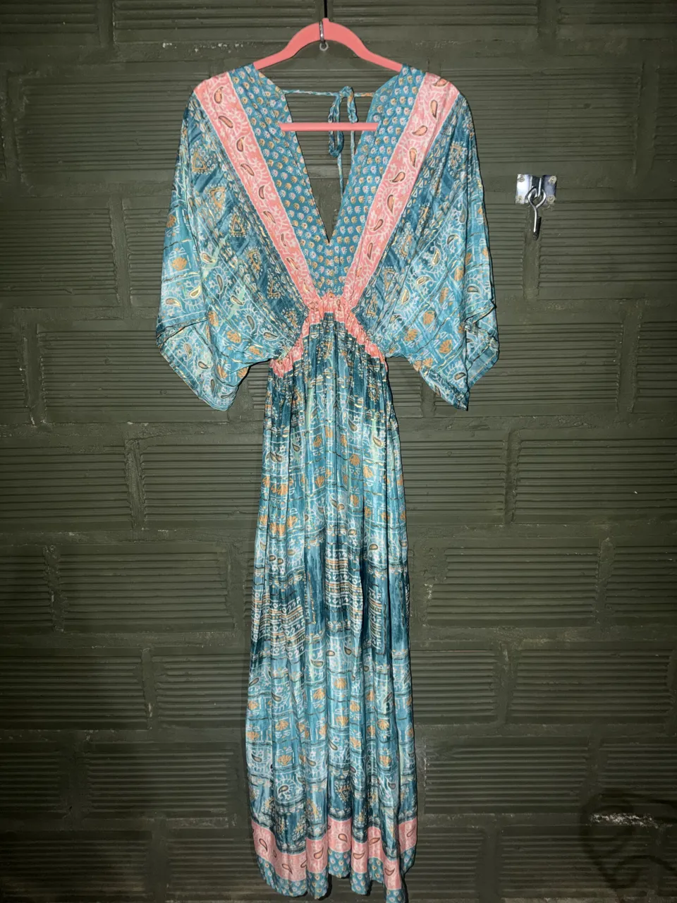 Vestido largo estampado en tonos verde y coral , con escote en V profundo y mangas amplias tipo murciélago. Posee elástico en la cintura y detalles de volados en el escote y la falda. La etiqueta indica "EL PLACARD DE FRAN".