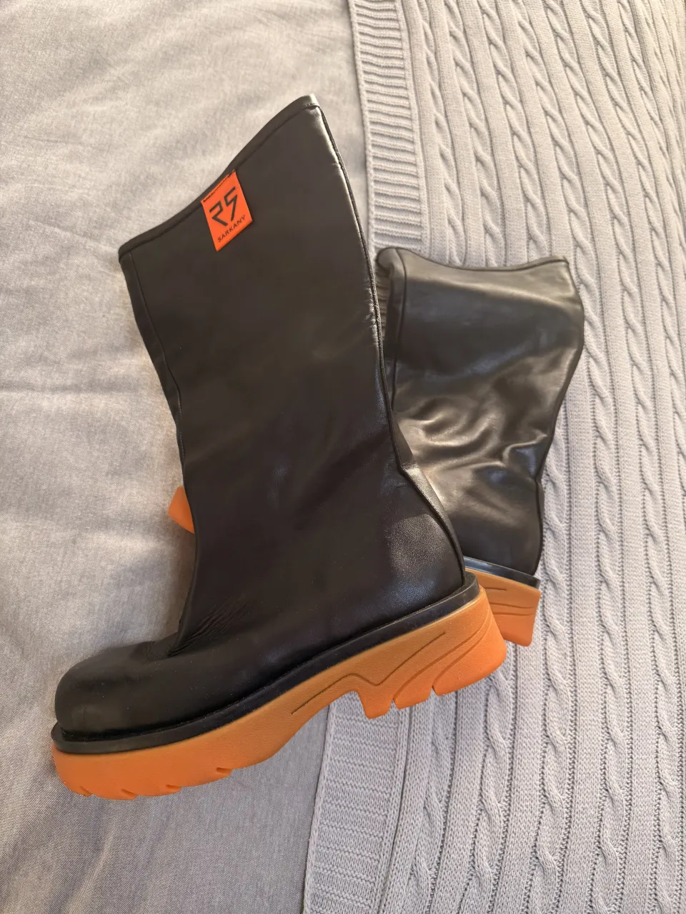 Botas negras con suela y detalles naranjas. Tienen una etiqueta naranja con el logo de la marca en el lateral.