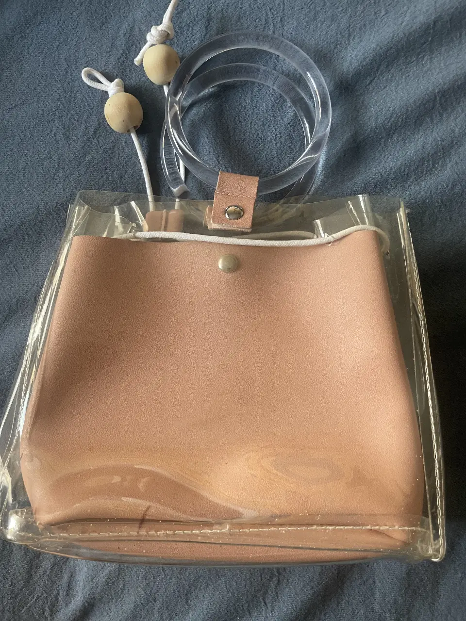 Cartera transparente con detalles en rosa pastel. Posee manijas de acrílico transparente y detalles de cuero sintético. Ideal para un look moderno y funcional.