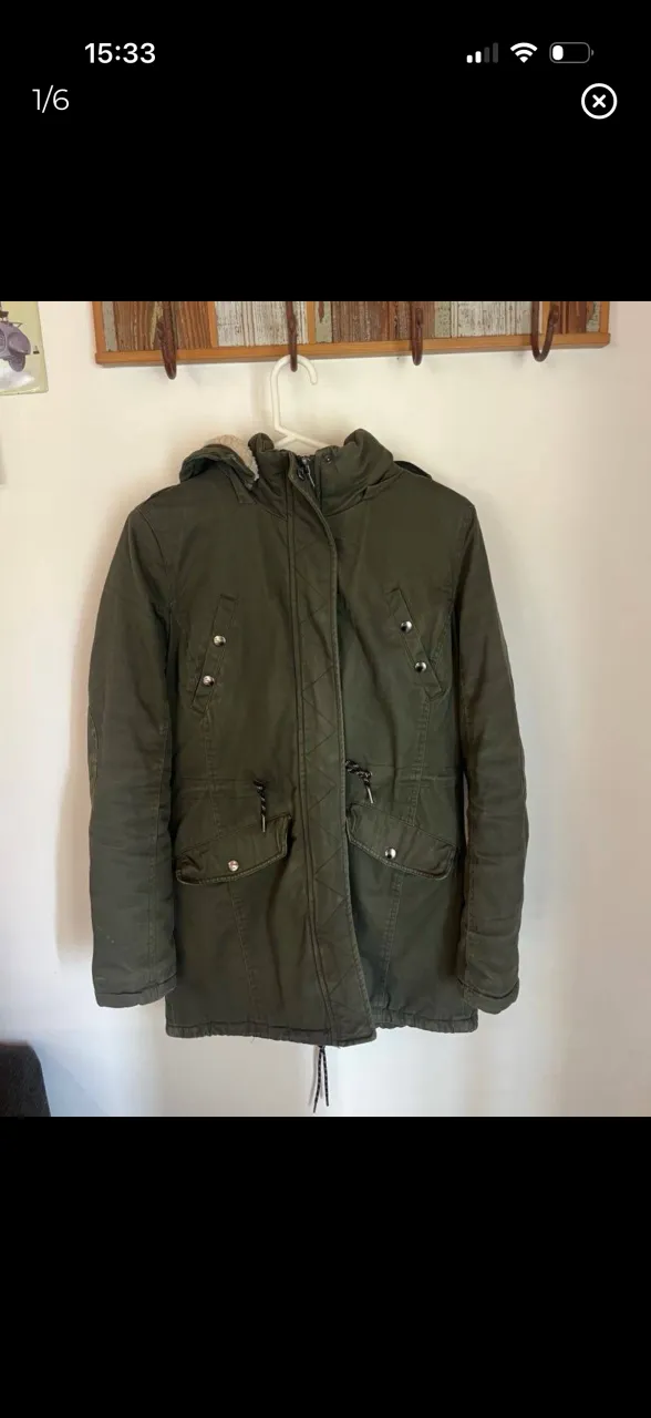 Parka verde con capucha forrada en corderito. Cierre principal con solapa y botones. Dos bolsillos frontales con tapa y cordón ajustable en la cintura. NO ES ZARA