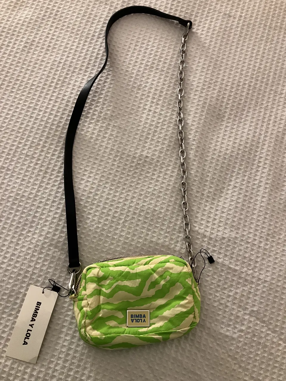 Cartera bandolera de la marca Bimba y Lola, con estampado de cebra en tonos verde y blanco. Cuenta con una correa negra y una cadena plateada para llevar al hombro. Incluye etiqueta y bolsa protectora.